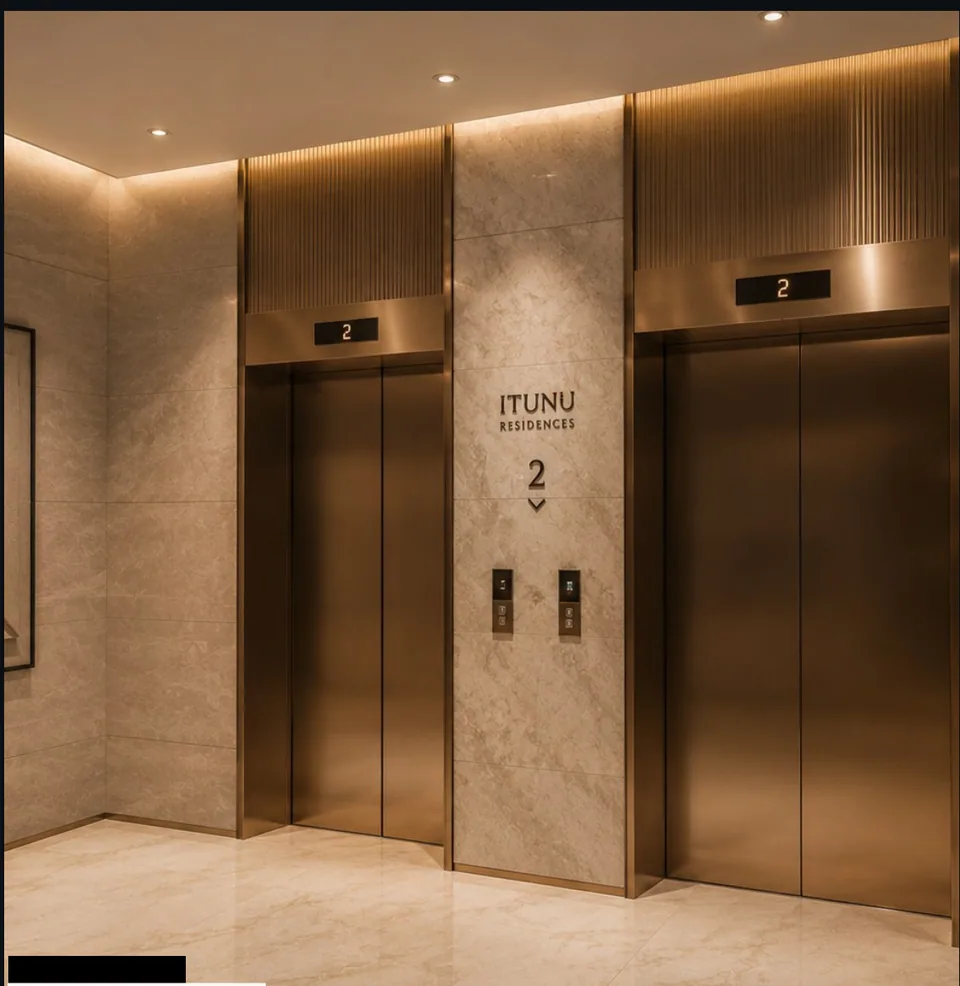 Lift lobby with two brushed-bronze passenger lift doors, floor number '2' in relief on a stone-clad pier, fluted bronze wall panels, framed artwork, large-format stone floor.