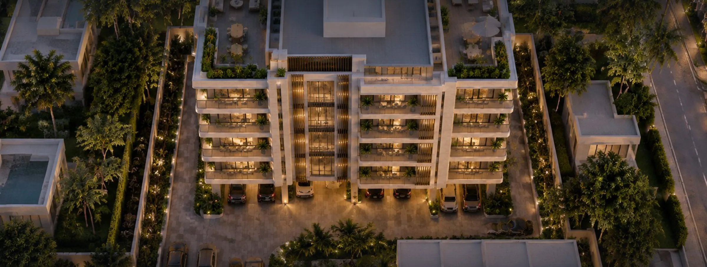 High aerial of the whole site in the evening: rooftop terrace with pergola, lounge seating and dining sets, the four residential floors, cars parked in the open ground-level undercroft, landscaped boundary, and the generator house and water treatment plant along the rear boundary.